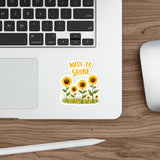 Laptop with a 'Made to Shine' sticker featuring sunflowers on a wooden desk.